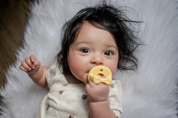 itzy ritzy sweetie soother™ pacifier sets (2 pack): aquamarine + peach bows Baby chewing on a yellow teething toy.