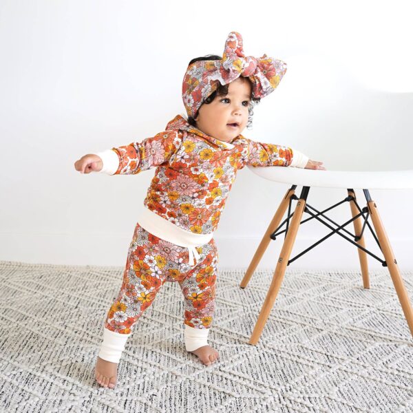 Baby in floral outfit standing by chair.