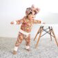 Baby in floral outfit standing by chair.