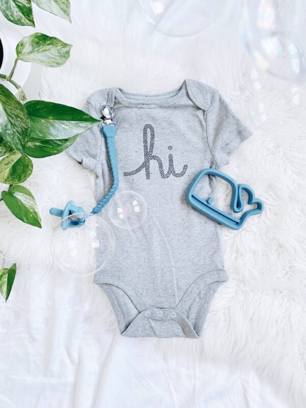 itzy ritzy sweetie soother™ pacifier sets (2 pack): agave + succulent cables Gray baby onesie with 'hi' text and baby toys.