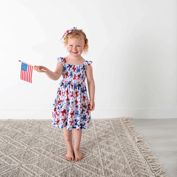 gigi and max eleanor bow clip bow 4th of july hair accessory Child holding American flag, wearing star-patterned dress.