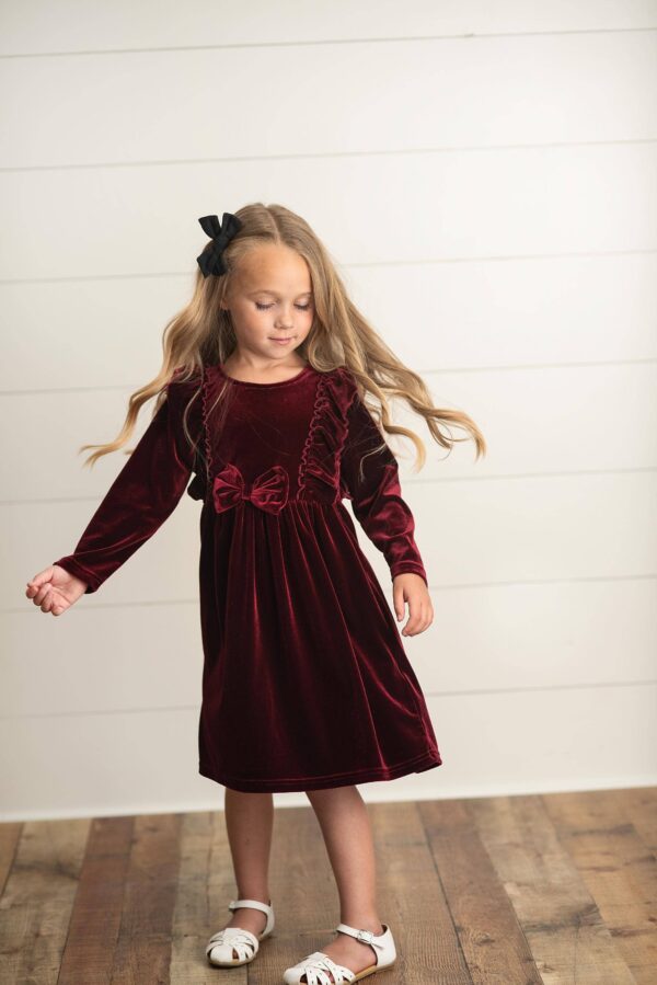 Child in red velvet dress and black bow standing.