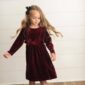 Child in red velvet dress and black bow standing.