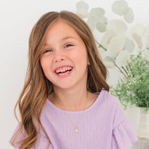Smiling child in purple dress, green plant background.