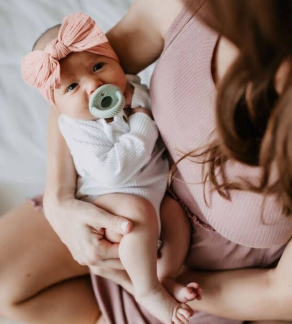 itzy ritzy sweetie soother™ pacifier sets (2 pack): agave + succulent cables Baby with pacifier and bow, held by woman.