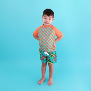 Child in checkered shirt with dinosaur shorts smiling.