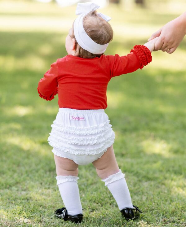 Baby in red shirt holding adult's hand outdoors.