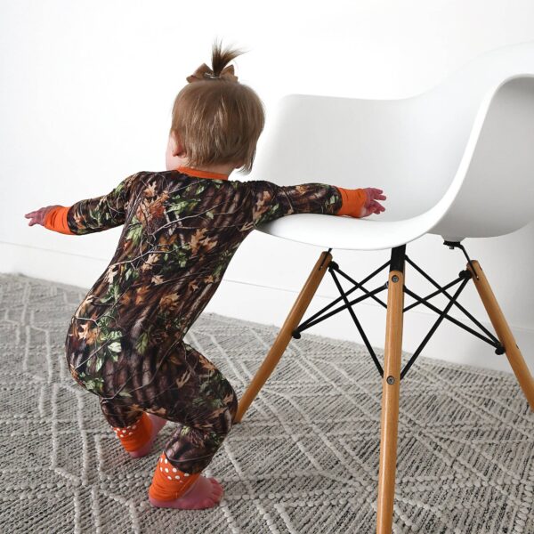 gigi and max hunter camo zip sleepies little bamboo baby outfit Toddler in camouflage pajamas leans on white chair.