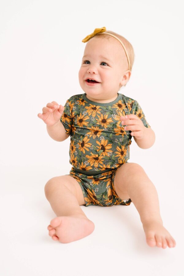 little one shop sunflower meadow bamboo shorts set Smiling baby in floral onesie with headband.