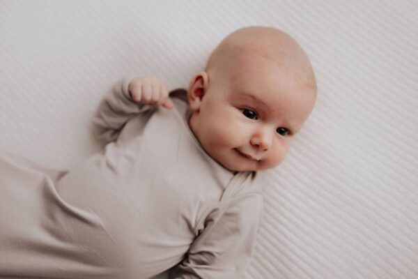 three little tots harbor mist taupe knotted gown Baby lying on white textured blanket, smiling.
