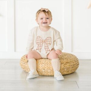 Smiling baby in bow outfit sitting on cushion.