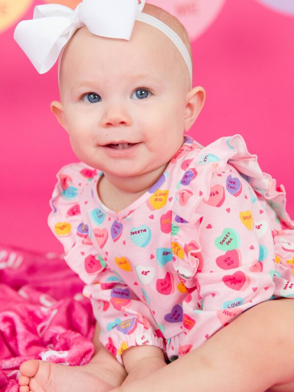rufflebutts + ruggedbutts baby girls be my valentine knit long sleeve waterfall bubble romper Baby wearing heart-print romper and white bow.