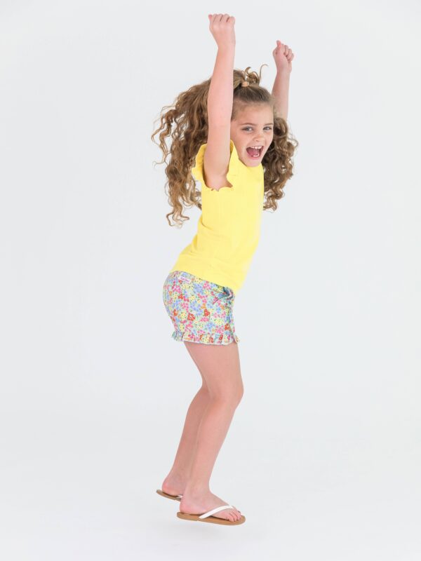 rufflebutts girls petaling around ruffle trim woven shorts and yellow ribbded top set Child jumping in yellow shirt and floral shorts
