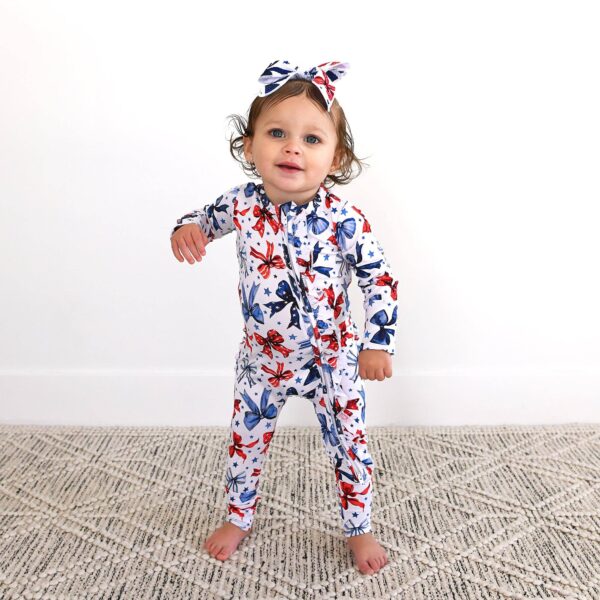 gigi and max eleanor bow clip bow 4th of july hair accessory Toddler in floral onesie and headbow stands smiling.