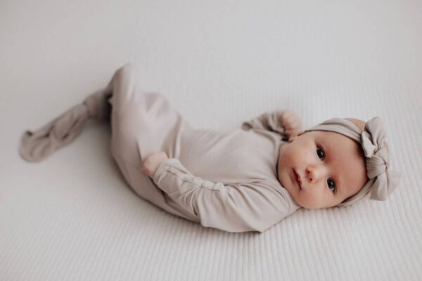three little tots harbor mist taupe knotted gown Baby in beige outfit and headband lying down.