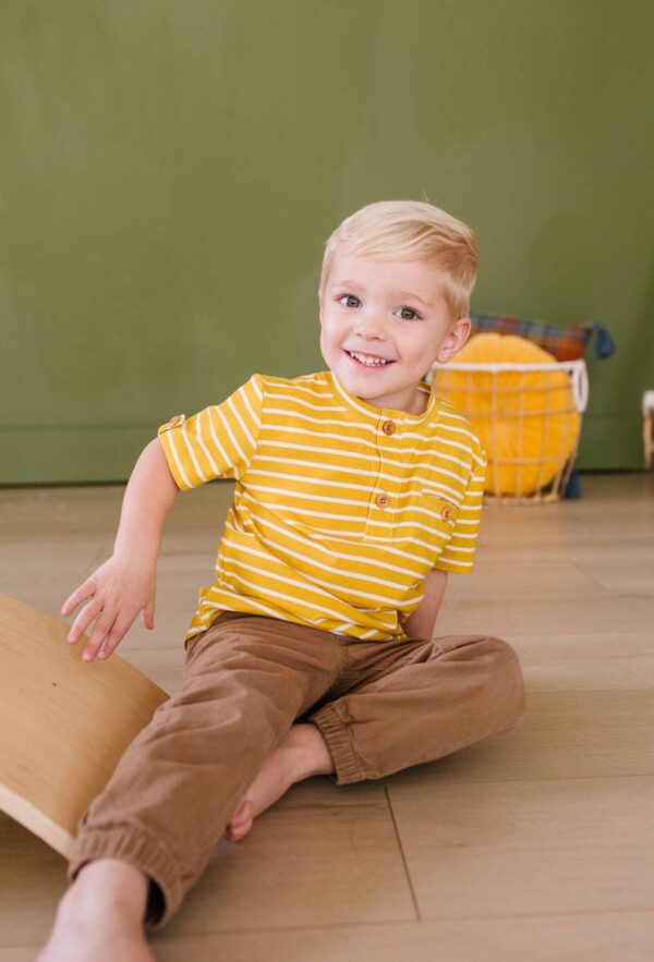 ollie jay alex tee in mustard stripe Smiling child in yellow striped shirt sitting indoors.
