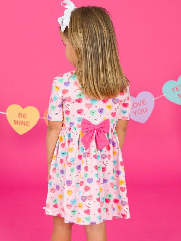 rufflebutts + ruggedbutts girls be my valentine short sleeve knit twirl dress Girl in heart-print dress with bow, pink background.