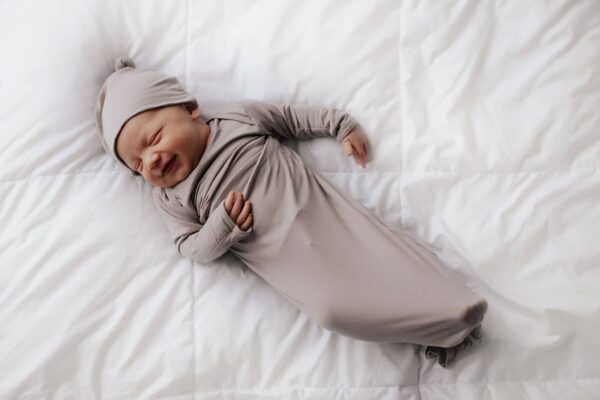 three little tots harbor mist taupe knotted gown Smiling baby in gray swaddle on bed