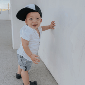 Smiling baby in cap and shorts against wall.