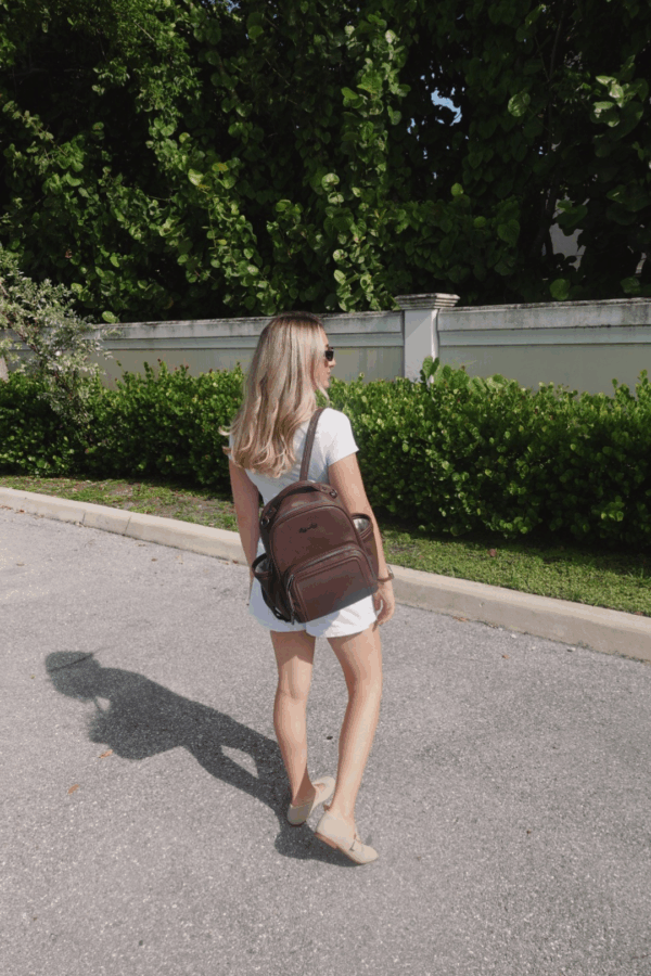 itzy ritzy *new* espresso itzy mini plus™ Woman wearing backpack walks on sunny sidewalk.