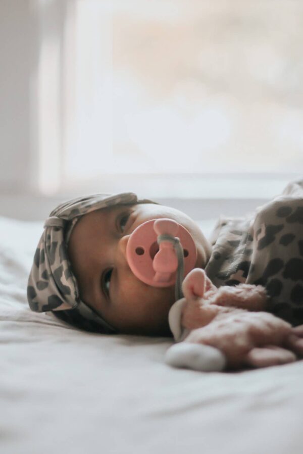 itzy ritzy sweetie soother™ pacifier sets (2 pack): agave + succulent cables Baby with pacifier and stuffed toy on bed.