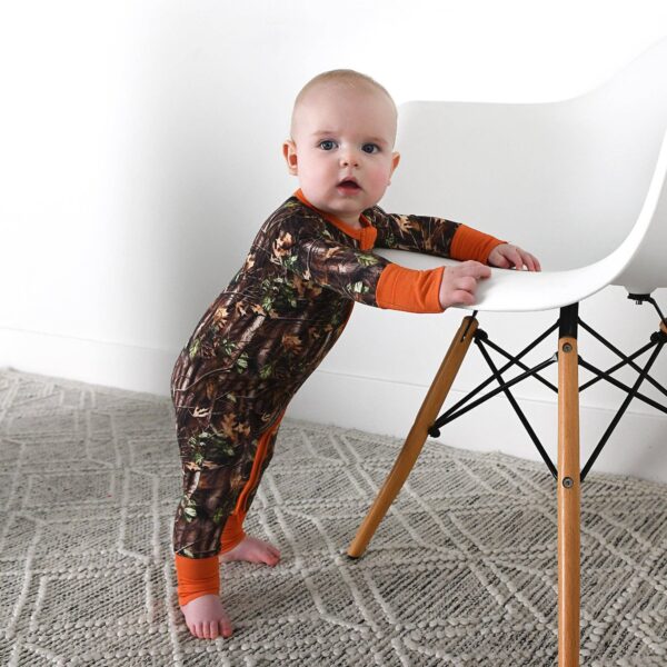 gigi and max hunter camo zip sleepies little bamboo baby outfit Baby in camouflage romper standing by chair.