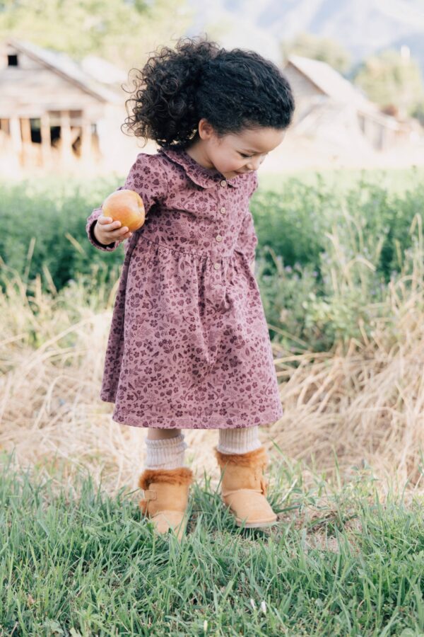 Child holding an apple outdoors, wearing patterned dress.