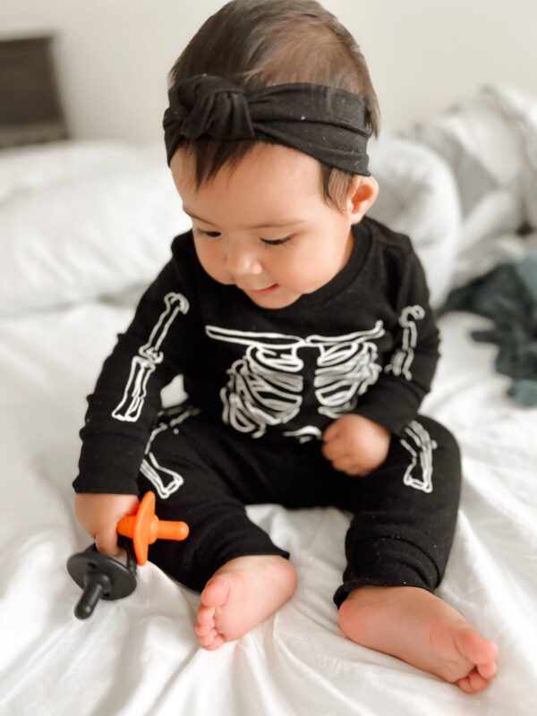 itzy ritzy sweetie soother™ pacifier sets (2 pack): aquamarine + peach bows Baby in skeleton outfit with pacifier on bed