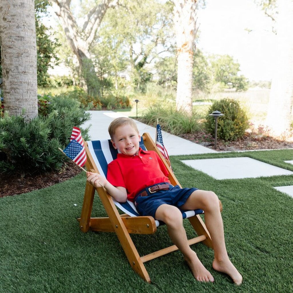 Young boy sitting outdoors in a striped deck chair holding two small American flags, wearing a bright red polo and navy twill shorts from a children’s boutique, patriotic summer outfit for kids