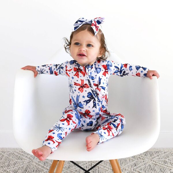 gigi and max eleanor bow clip bow 4th of july hair accessory Baby in floral outfit on white chair, smiling.