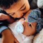 Child cuddling baby with pacifier and bow.