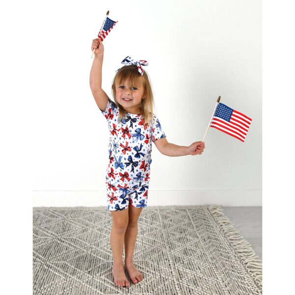 gigi and max eleanor bow clip bow 4th of july hair accessory Child waving American flags joyfully in printed outfit.