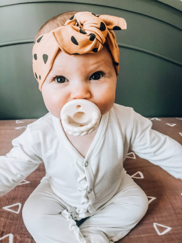 itzy ritzy sweetie soother™ pacifier sets (2 pack): agave + succulent cables Baby with headband and teether sitting on bed.