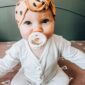 Baby with headband and teether sitting on bed.