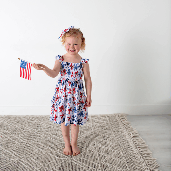 gigi and max eleanor bow clip bow 4th of july hair accessory Smiling child holding American flag indoors.