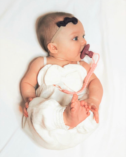 itzy ritzy sweetie soother™ pacifier sets (2 pack): mustard + gray braids Baby with pacifier in white outfit on bed.