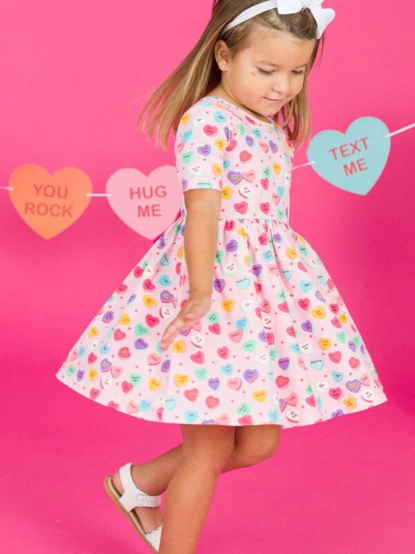 rufflebutts + ruggedbutts girls be my valentine short sleeve knit twirl dress Child in dress with heart pattern, pink background.