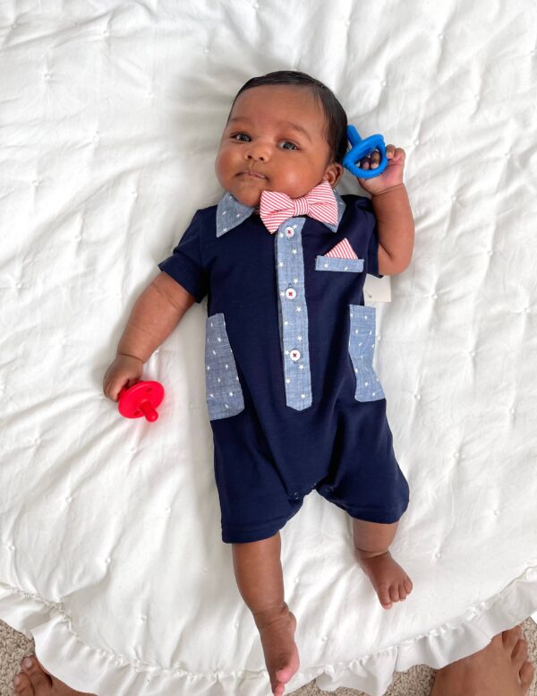 itzy ritzy sweetie soother™ pacifier sets (2 pack): aquamarine + peach bows Baby in navy romper with pacifiers on bed.