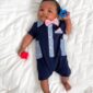 Baby in navy romper with pacifiers on bed.