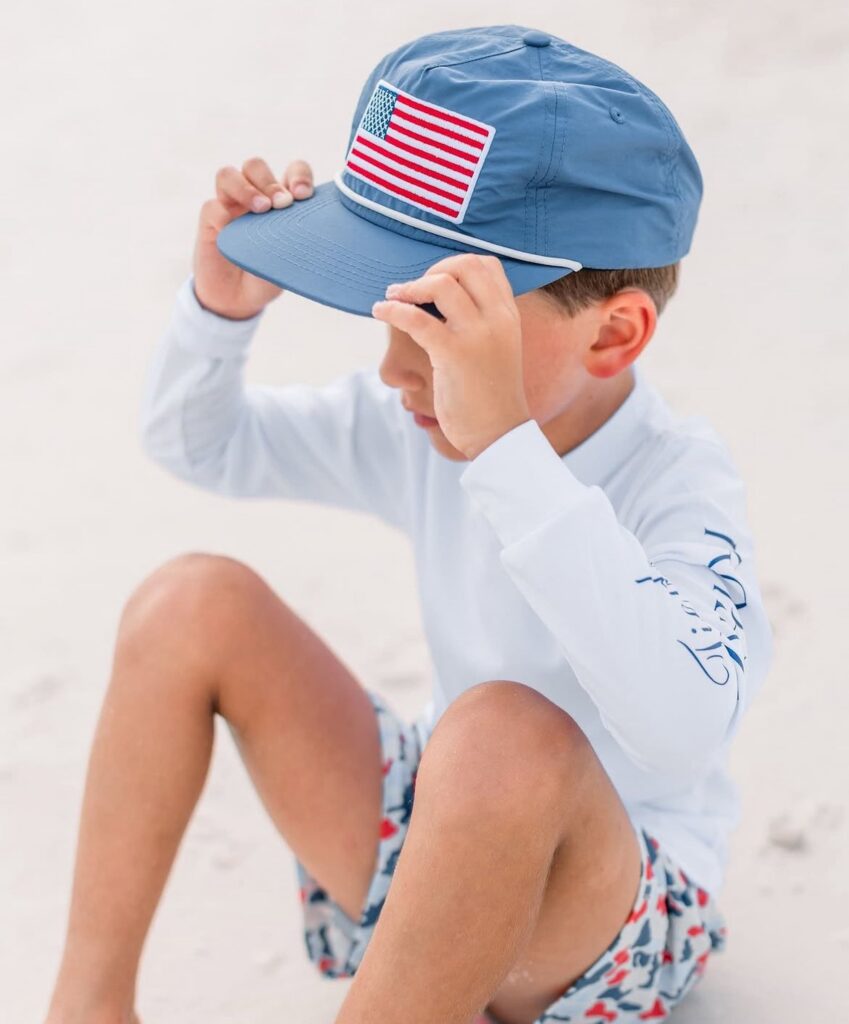 young boy wearing a blue hat with American flag patch, adjusting cap, patriotic kids accessory for summer, Properly Tied boys hat