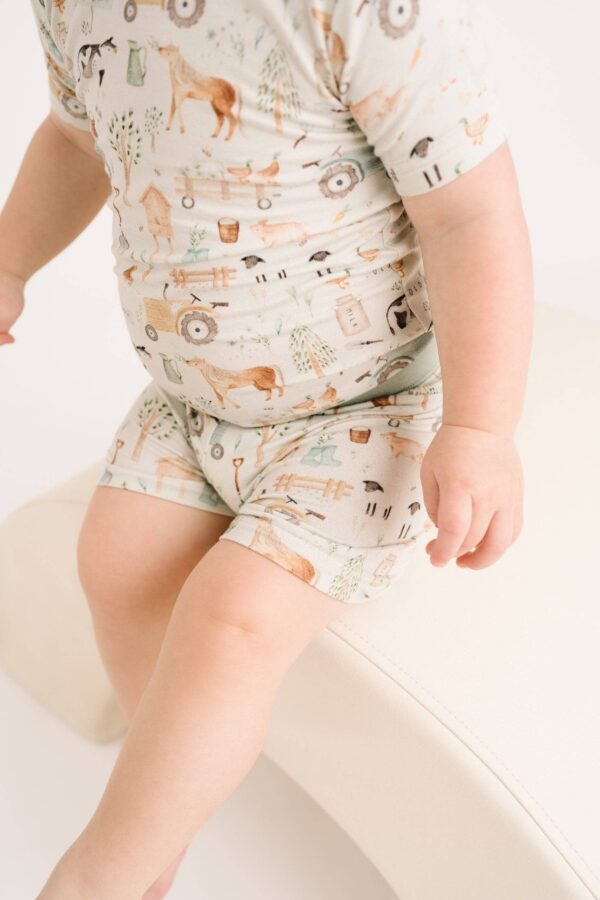 little one shop little farm bamboo shorts set Baby wearing farm-themed pajama set, sitting on white.