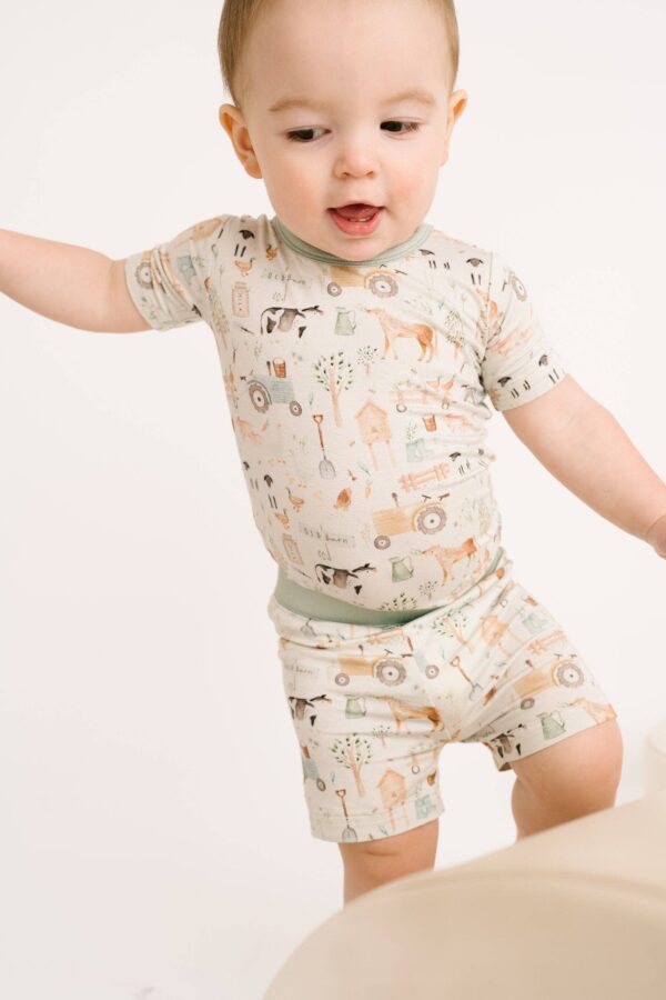 little one shop little farm bamboo shorts set Baby wearing farm-themed outfit, standing on one leg.