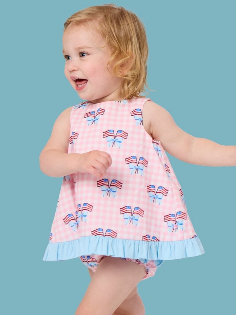Blonde headed little girl, blue background, wearing a pink gingham patriotic flag swing top and bloomers.