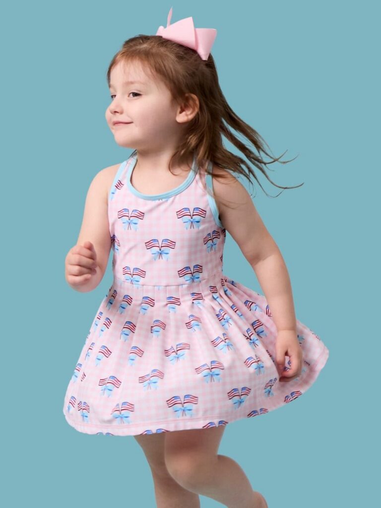 Brown headed girl on blue background wearing a pink gingham active tennis dress with flags.
