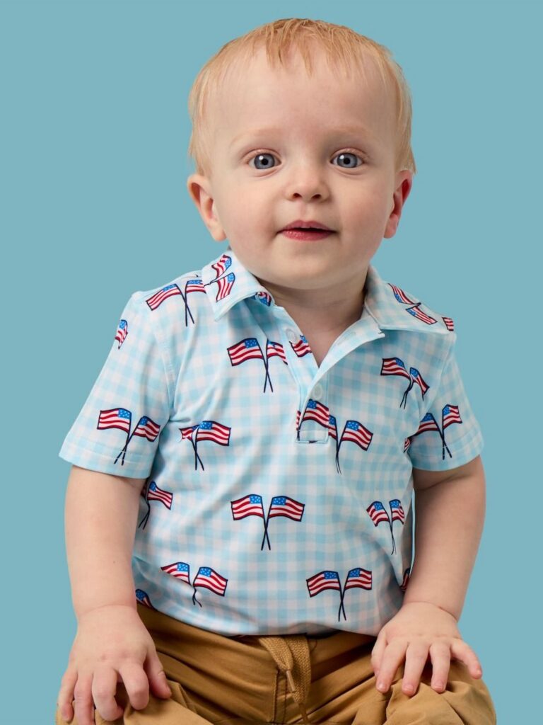 Baby boy on blue background wearing a baby blue gingham polo with flags.