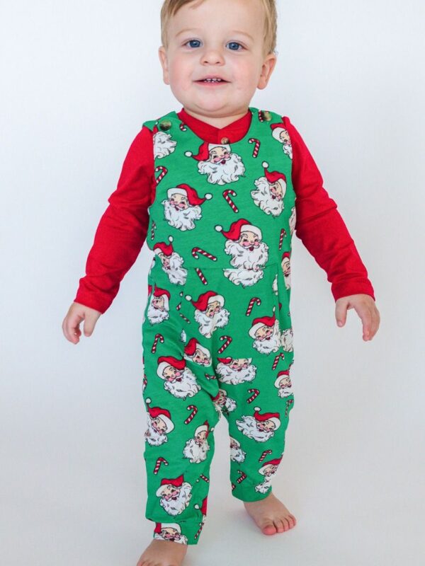 ruggedbutts green santa joy overalls and red henley onesie set Child in Christmas-themed pajamas smiles.
