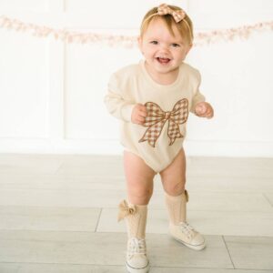 Smiling baby in bow outfit standing indoors.