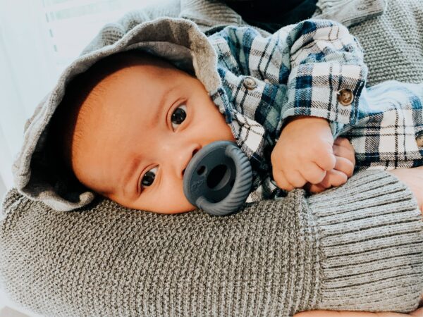 itzy ritzy sweetie soother™ pacifier sets (2 pack): agave + succulent cables Baby resting, holding gray pacifier, wearing plaid shirt.