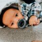 Baby resting, holding gray pacifier, wearing plaid shirt.
