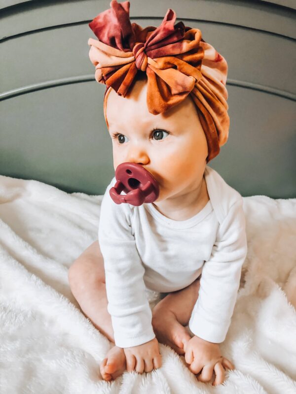 itzy ritzy sweetie soother™ pacifier sets (2 pack): aquamarine + peach bows Baby with headwrap and pacifier on blanket.